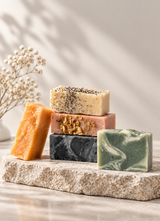 Calinea handmade soap bars stacked on travertine — olive, jasmine, charcoal and laurel cold-process bars